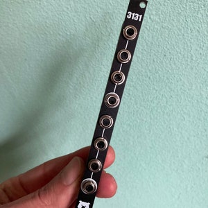 Könnte beinhalten: Ein schwarzes elektronisches Bauteil mit mehreren silbernen Buchsen und der weißen Aufschrift "3131". Das Bauteil hat ein kleines Bärenlogo und wird vor einem hellgrünen Hintergrund gehalten.