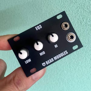 Puede incluir: Módulo de audio EQ3 negro con perillas blancas etiquetadas LOW, MID e HI. El módulo tiene dos conectores de entrada y el texto "BEAR MODULES". El módulo está sostenido por una mano.