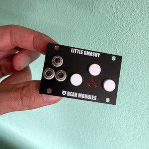 Puede incluir: Un módulo de música electrónica negro con el texto "LITTLE SMASHY" y "BEAR MODULES". El módulo tiene tres botones plateados, tres botones blancos y tres LED rojos.