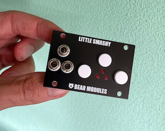 May include: A black electronic music module with the text "LITTLE SMASHY" and "BEAR MODULES". The module has three silver knobs, three white buttons, and three red LEDs.
