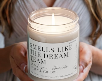 Może przedstawiać: Biała świeca sojowa z czarną etykietą, na której widnieje napis "Smells Like The Dream Team Thank you. Amanda FOR ALL YOU DO!" Świeca jest zapalona i płomień jest widoczny.