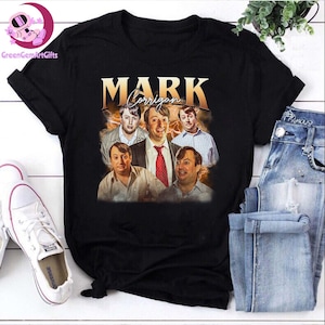 Peut inclure: T-shirt noir avec un graphisme de style vintage de Mark Corrigan de la sitcom britannique Peep Show. Le graphisme comprend plusieurs images du personnage et le nom "MARK Corrigan" en lettres dorées. Le t-shirt est présenté avec un jean bleu et des baskets blanches.