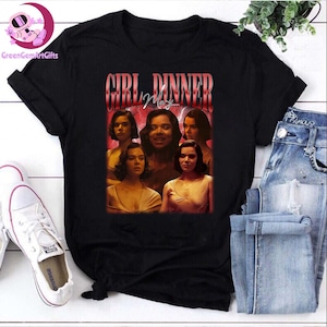 以下が含まれることがあります： 「GIRL DINNER」の文字が赤と白で描かれたレトロスタイルのグラフィックが特徴の黒いTシャツ。女性のコラージュ画像がプリントされています。青いジーンズと白いスニーカーと組み合わせています。