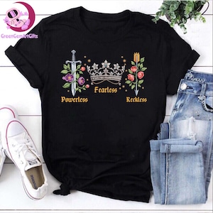 May include: Black t-shirt with a graphic design featuring a sword, crown, and arrow with flowers and the words "Powerless", "Fearless", and "Reckless".
