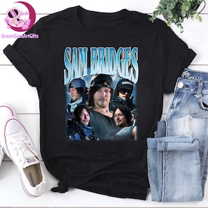 Può includere: T-shirt nero con un collage di immagini di Sam Bridges e la scritta "SAM BRIDGES" in blu e bianco. La maglietta è abbinata a jeans blu e scarpe da ginnastica bianche.