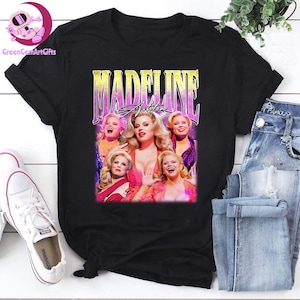 May include: Black t-shirt featuring a graphic of a woman with blonde hair and the text "MADELINE Ashton". The graphic has a vintage, retro style with a pink and purple gradient. The shirt is paired with blue jeans and white sneakers.