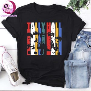 Może przedstawiać: Czarny t-shirt z nazwą zespołu "TALLY HALL" nad kolorową grafiką członków zespołu. Grafika wykorzystuje pionowe paski w kolorach czerwonym, żółtym, niebieskim i białym. Widoczne są białe trampki i niebieskie dżinsy.
