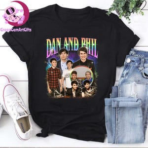 May include: Black t-shirt featuring a retro-style graphic of multiple people with the text "DAN AND PHIL" in a colorful, stylized font. The design includes a collage of images. The shirt is displayed with jeans and a pair of white sneakers.