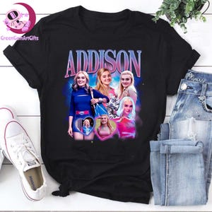 May include: Black t-shirt with the name "ADDISON" in a retro style above a collage of images. The collage includes multiple photos of a person in different outfits. The shirt is displayed with blue jeans and white trainers.