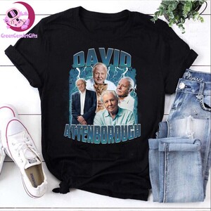 Op de afbeelding: Zwart t-shirt met een afbeelding van David Attenborough en de tekst "DAVID ATTENBOROUGH" in blauw en wit. Het ontwerp bevat meerdere afbeeldingen van Attenborough. Het shirt wordt getoond met een blauwe jeans en witte sneakers.