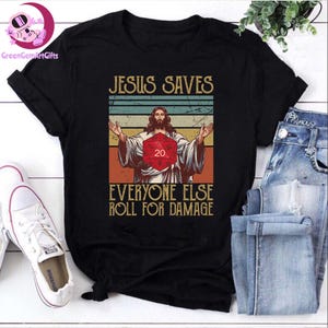 May include: A black t-shirt with a vintage-style graphic of Jesus holding a twenty-sided die. The text reads "Jesus Saves Everyone Else Roll For Damage".