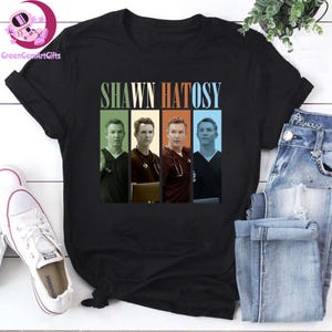 Könnte beinhalten: Schwarzes T-Shirt mit dem Namen "SHAWN HATOSY" über vier Bildern des Schauspielers in vertikalen Farbblöcken. Die Blöcke sind grün, braun, orange und blau. Weiße Turnschuhe und blaue Jeans sind im Hintergrund.
