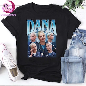 May include: Black t-shirt featuring a graphic of a woman with the name "DANA Evans" in a blue, vintage-style font. The graphic includes multiple images of the woman in a medical setting. A pair of white sneakers and blue jeans are visible.