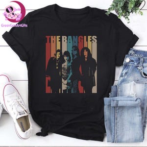 May include: Black t-shirt featuring the band "The Bangles" with a retro design. The design includes a vertical striped background in red, orange, beige, blue, and brown. The band members are silhouetted in the design.