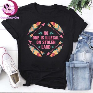 May include: Black t-shirt with a pink and blue graphic design of arrows and feathers. The text "NO ONE IS ILLEGAL ON STOLEN LAND" is written in pink.