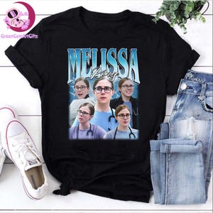 May include: Black t-shirt with a graphic design featuring the name "Melissa R." in a blue, vintage-style font. The design includes multiple images of a person wearing glasses and a stethoscope. The shirt is paired with blue jeans and white trainers.