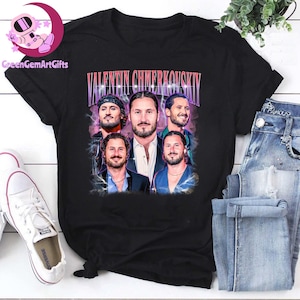 May include: Black t-shirt featuring a vintage-style graphic of Valentin Chmerkovskiy. The design includes multiple images of the dancer's face and the name "VALENTIN CHMERKOVSKIY" in a retro font. The shirt is paired with blue jeans and white sneakers.