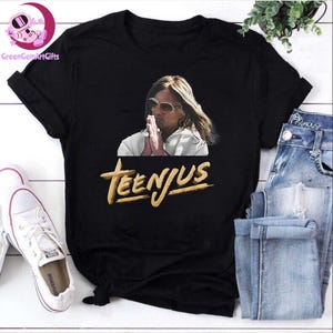 Pode incluir: Camiseta preta com um gráfico de uma pessoa usando óculos de sol e mãos juntas. O texto dourado "TEENJUS" é impresso abaixo. A camiseta é combinada com jeans azuis e tênis brancos.