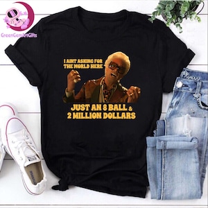 Pode incluir: Camiseta preta com um gráfico de um homem com óculos de sol e o texto "I AINT ASKING FOR THE WORLD HERE JUST AN 8 BALL & 2 MILLION DOLLARS." A camiseta é combinada com jeans azuis e tênis brancos.