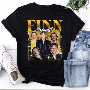 Könnte beinhalten: Ein schwarzes Kurzarm-T-Shirt mit einem Grafikdruck von Finn Bennett. Das Design zeigt den Namen FINN BENNETT in großen gelben und weißen Buchstaben über einer Collage aus fünf fotografischen Porträts derselben Person, akzentuiert durch gelben Rauch.