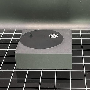 May include: A black circular display stand with a small black pin in the center and a white "DC" logo. The stand is made of a textured material and has a square base.