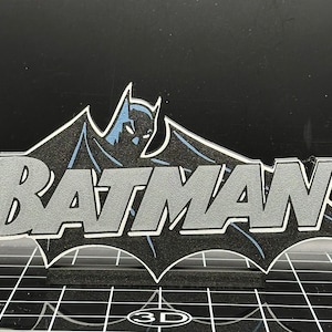 May include: A black and silver 3D wall decor featuring the Batman logo with the text "BATMAN" in a bold, stylized font.