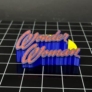 May include: A blue and red 3D-printed Wonder Woman logo. The text "Wonder Woman" is in a stylized script with a red glitter effect, set against a blue, layered background. The logo is on a black surface.