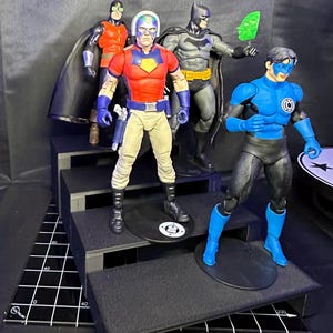 May include: Four action figures of comic book characters are displayed on a tiered platform. The figures include Batman, a character in a red and blue outfit, a character in a blue and black outfit, and a character in an orange and black outfit.
