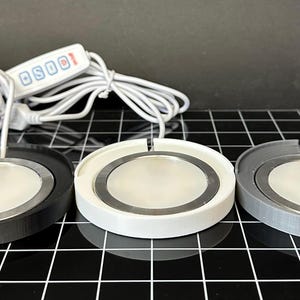 May include: Three circular LED light panels in black, white, and gray, each with a silver center and a white cord. A white control panel with the text "OS001" is visible. The lights are on a black grid surface.