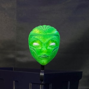 Puede incluir: Una máscara alienígena verde con ojos blancos y rasgos faciales detallados. La máscara es de color verde vibrante y parece estar impresa en 3D. La máscara está montada sobre un soporte negro, sobre un fondo oscuro.