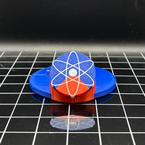 May include: A blue and red 3D printed atom symbol on a blue base. The atom symbol is white with a red center and a white circle in the center.
