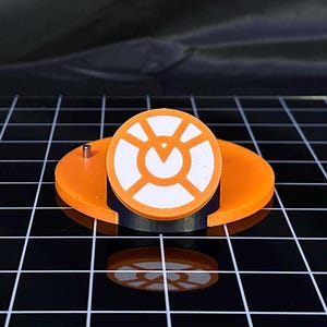 Puede incluir: Un objeto circular naranja y blanco con un diseño geométrico, montado sobre una base negra y un disco naranja. Tiene un pequeño pasador metálico. El diseño es simétrico, probablemente un elemento decorativo.