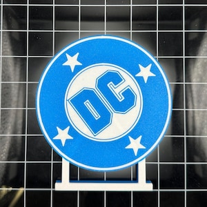 Puede incluir: Un logotipo de DC Comics impreso en 3D azul y blanco con cinco estrellas blancas alrededor del círculo. El logotipo está en un soporte blanco.
