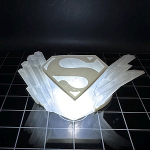 Pode incluir: Um logotipo do Superman branco impresso em 3D com uma base cristalina branca. O logotipo é iluminado por dentro, projetando um brilho quente.