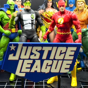 Puede incluir: Un letrero azul y blanco de la Liga de la Justicia con una estrella en la esquina superior izquierda. El letrero está sostenido por un soporte negro. El letrero está frente a un grupo de figuras de acción, que incluyen Superman, Wonder Woman, Flash, Linterna Verde, Aquaman y Martian Manhunter.
