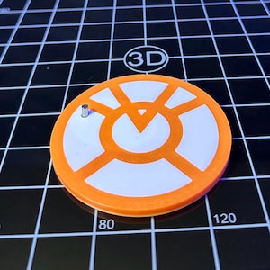 Puede incluir: Un objeto circular naranja y blanco con un pequeño pasador de metal. El objeto tiene un diseño geométrico con un punto central y líneas radiales. El fondo es una cuadrícula negra con los números 80 y 120 visibles.