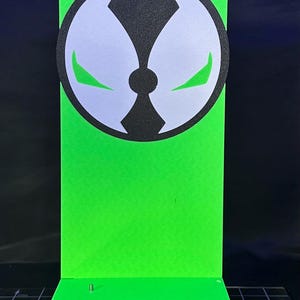 May include: A neon green display stand featuring the Spawn logo. The logo is a white and black circular design with green accents. The stand has a rectangular base and a vertical back panel, perfect for displaying collectibles.