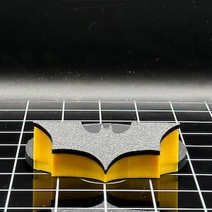 May include: A black and yellow 3D printed Batman logo. The logo is on a black surface.