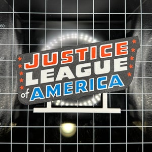 Puede incluir: Un letrero blanco con las palabras "Justice League of America" en letras rojas, blancas y azules. El letrero está en un soporte blanco.