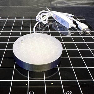 May include: A round, light-colored platform with a dark rim, connected to a white USB cable and control panel. The platform has a small, cylindrical protrusion on top. The control panel has four buttons. The platform is on a black grid.