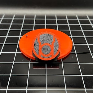 Puede incluir: Soporte impreso en 3D de color naranja y negro con un símbolo negro y naranja. El símbolo es un círculo con un anillo alrededor y una línea que lo atraviesa por el centro.