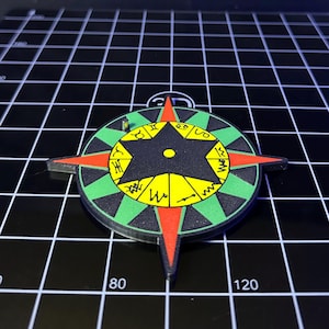 May include: A circular pendant with a compass rose design. The pendant features a black star in the center, surrounded by a yellow and green ring with zodiac symbols. Red and black compass points extend from the circle.