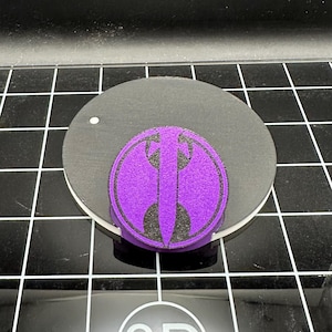 May include: A purple round sticker with a black symbol of a stylised sword with wings. The sticker is on a black surface.