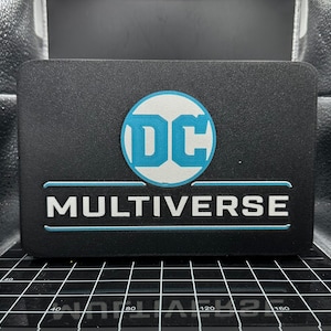 May include: A black rectangular box with a white DC logo inside a blue circle and the text "MULTIVERSE" in white letters with a blue outline.