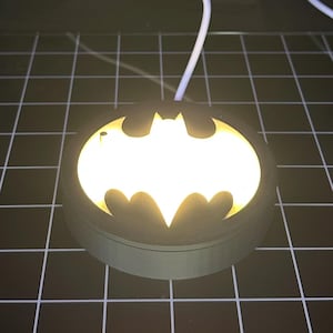 May include: A glowing Batman symbol night light. The light is shaped like the iconic bat symbol, emitting a bright, warm light. The light is set in a dark, circular base, with a white cord extending from the top.
