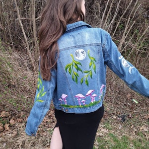 May include: A light blue denim jacket with hand-painted artwork. The back features a full moon, green leaves, and purple mushrooms. The sleeves have additional leaf and moon phase designs. A unique, artistic piece.