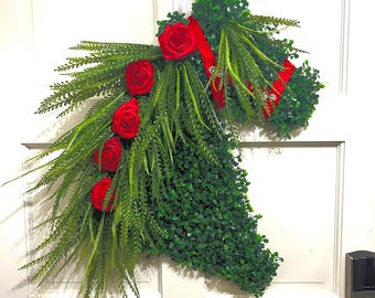 May include: A horse head-shaped wreath constructed from dense green artificial boxwood foliage. The mane is composed of long, feathery green fern-like fronds accented with five vibrant red roses and a bright red ribbon strap across the neck.