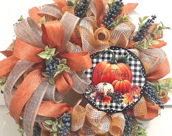 May include: A decorative wreath featuring orange and tan mesh ribbons, dark blue berry sprigs, and green leaves. A circular sign in the center displays a black and white buffalo check pattern with orange pumpkins and the text Give Thanks.