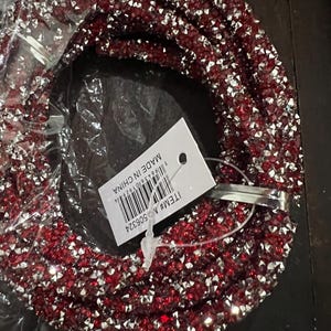 May include: A red and silver beaded necklace, partially visible within its packaging. The beads are small and closely packed, creating a sparkling effect. A tag attached to the necklace reads "MADE IN CHINA" and includes a barcode and item number.