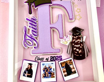 May include: A white shadow box with a purple letter F, a graduation cap, and the words "You did it!" and "Class of 2025". There are three photos in the shadow box, one of a graduate in a cap and gown, one of a group of graduates, and one of a graduate in a cap and gown walking away from the camera.